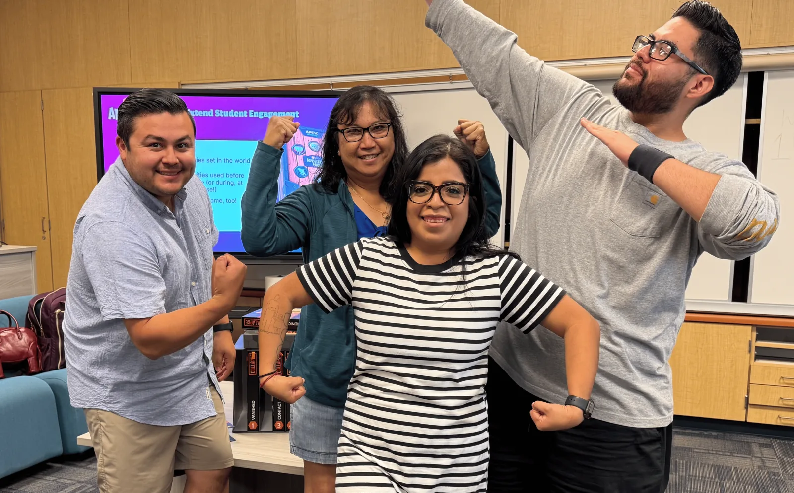 Four educators strike playful flexing poses together in front of a training screen